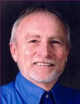 A smiling older man with gray hair and a beard, wearing a blue button-down shirt.